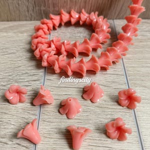 May include: A string of coral pink flower-shaped beads arranged in a circular pattern, accompanied by several loose individual flower beads. The text E# Coral Pink and findingscity are visible in the image. Each bead features a central hole.