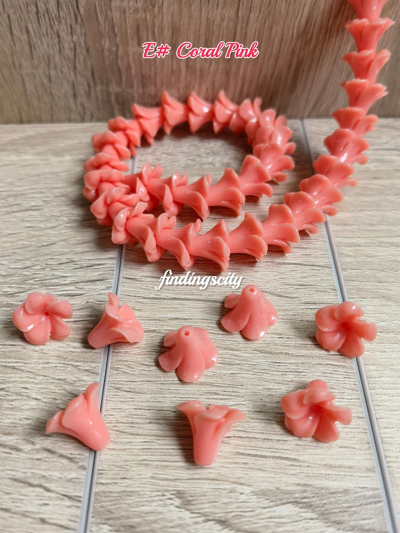 May include: A string of coral pink flower-shaped beads arranged in a circular pattern, accompanied by several loose individual flower beads. The text E# Coral Pink and findingscity are visible in the image. Each bead features a central hole.
