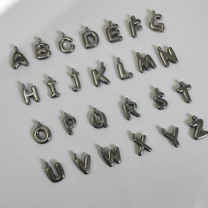 May include: A set of 26 silver-coloured metal alphabet letter charms. Each letter is in a bubble-like font style.