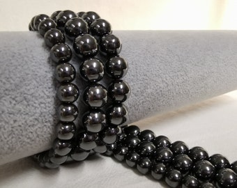 May include: A strand of black hematite beads, with a high polish finish, arranged in a circular pattern on a gray surface.