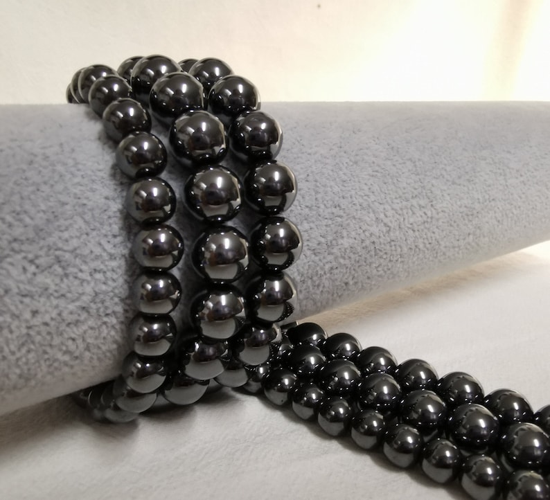 May include: A strand of black hematite beads, with a high polish finish, arranged in a circular pattern on a gray surface.