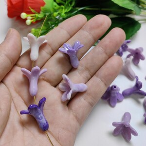 May include: A collection of small, flower-shaped beads in shades of purple and white. The beads are strung on a thin gold wire, and are likely for jewelry making or other crafts. The beads have a glossy finish and are clustered together.