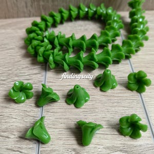 May include: A collection of green flower-shaped beads, including a long strand coiled in a circle and several loose individual beads. Each bead features a five-petal design with a central hole for stringing. The text A# Green and findingscity appear.
