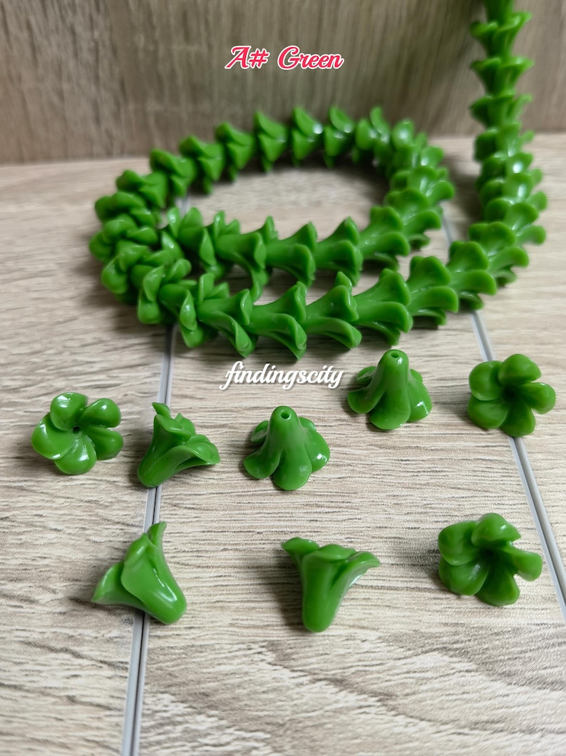 May include: A collection of green flower-shaped beads, including a long strand coiled in a circle and several loose individual beads. Each bead features a five-petal design with a central hole for stringing. The text A# Green and findingscity appear.