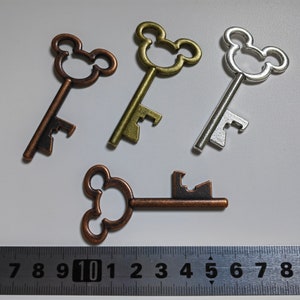 Puede incluir: Cuatro llaves decorativas con diseño de cabeza de Mickey Mouse. Las llaves son de tonos bronce, latón y plata. Una llave está plana. Una regla en la parte inferior muestra las medidas en centímetros.