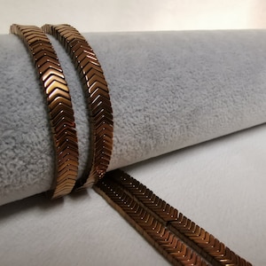 May include: Two strands of brown chevron-shaped beads, lying on a white surface. The beads are made of metal and have a shiny finish.