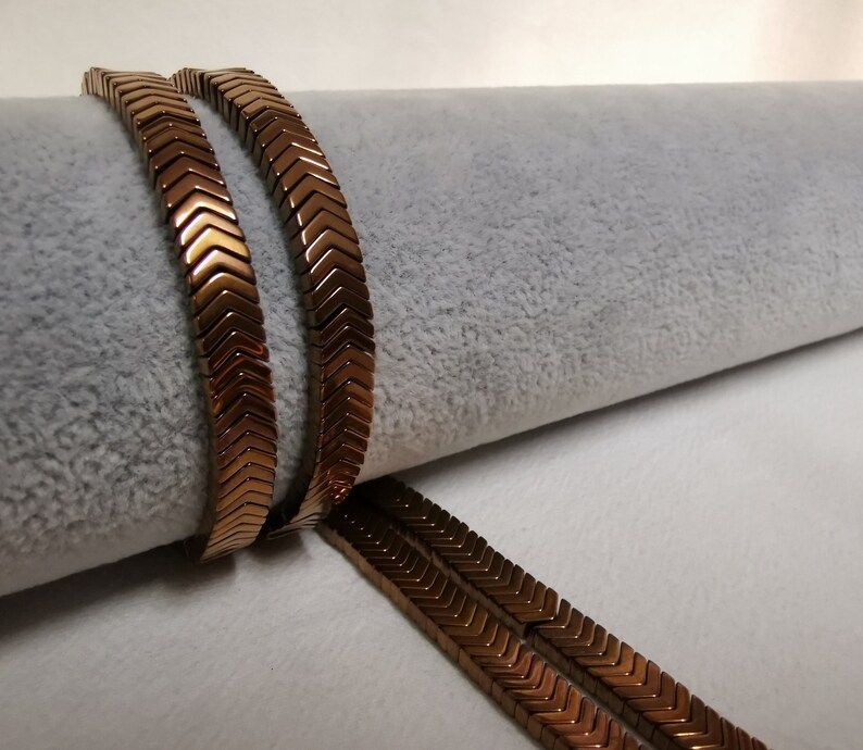 May include: Two strands of brown chevron-shaped beads, lying on a white surface. The beads are made of metal and have a shiny finish.