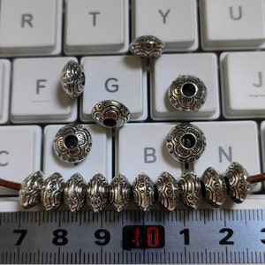 May include: A close-up of ten silver metal beads with a decorative floral design. The beads are arranged in a row on a ruler with the numbers 6 to 10 visible.