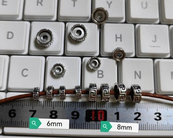 May include: A close-up of silver metal beads with a Greek key design. The beads are 6mm and 8mm in diameter. The beads are on a brown leather cord and are being measured with a ruler.