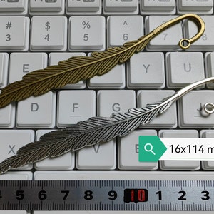 May include: Two feather-shaped bookmarks, one gold and one silver, with a curled tip. The bookmarks are laid out on a white keyboard with a ruler measuring 16x114 mm.