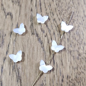 May include: Six white mother-of-pearl butterfly charms on a wooden surface. The charms are small and delicate, with a natural iridescent sheen.