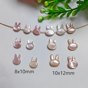 Op de afbeelding: Assortiment van roze en witte schelpjes bunny charms in twee maten: 8x10mm en 10x12mm. De charms zijn geregen aan een gouden draad.