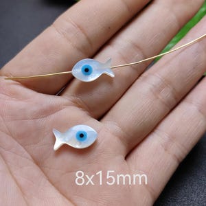 Może przedstawiać: Dwa małe zawieszki w kształcie rybki z perłowym wykończeniem i niebieskim wzorem oka. Zawieszki mają wymiary około 8 mm x 15 mm. Przez zawieszki przechodzi cienki, złoty drut. Obraz zawiera tekst "8x15mm".