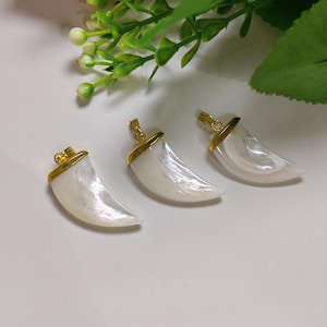May include: Three pearlescent, crescent-shaped pendants with gold-colored metal accents. The pendants are arranged on a white surface, with green foliage in the background. The pendants are likely for jewelry making or as decorative elements.