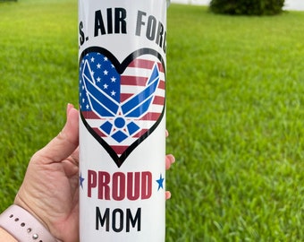 Może przedstawiać: Biały kubek z czarną pokrywką z napisem "U.S. AIR FORCE" i "PROUD MOM". Projekt zawiera grafikę w kształcie serca z amerykańską flagą i emblematem Sił Powietrznych. Niebieskie gwiazdy akcentują tekst.