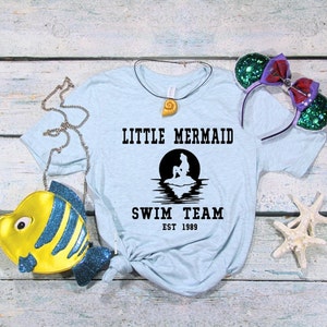 Puede incluir: Camiseta azul claro con una silueta negra de una sirena sentada en una roca con una luna llena detr&aacute;s de ella. El texto "Little Mermaid Swim Team Est 1989" est&aacute; impreso en la camiseta.