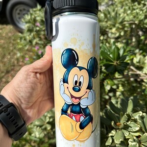 May include: A white stainless steel water bottle with a black lid and handle. The bottle has a colorful image of Mickey Mouse on it. The image is a watercolor style with yellow, orange, and blue colors.