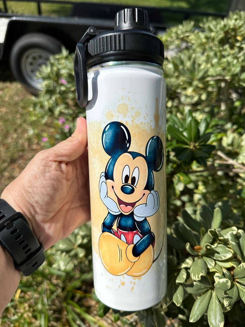 May include: A white stainless steel water bottle with a black lid and handle. The bottle has a colorful image of Mickey Mouse on it. The image is a watercolor style with yellow, orange, and blue colors.