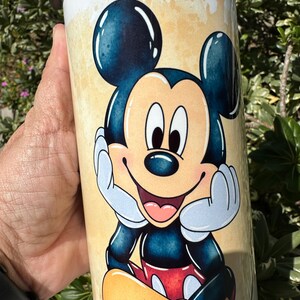 May include: A white tumbler with a cartoon image of Mickey Mouse. Mickey is smiling and has his hands on his chin. The background is a light yellow with brown splatters.
