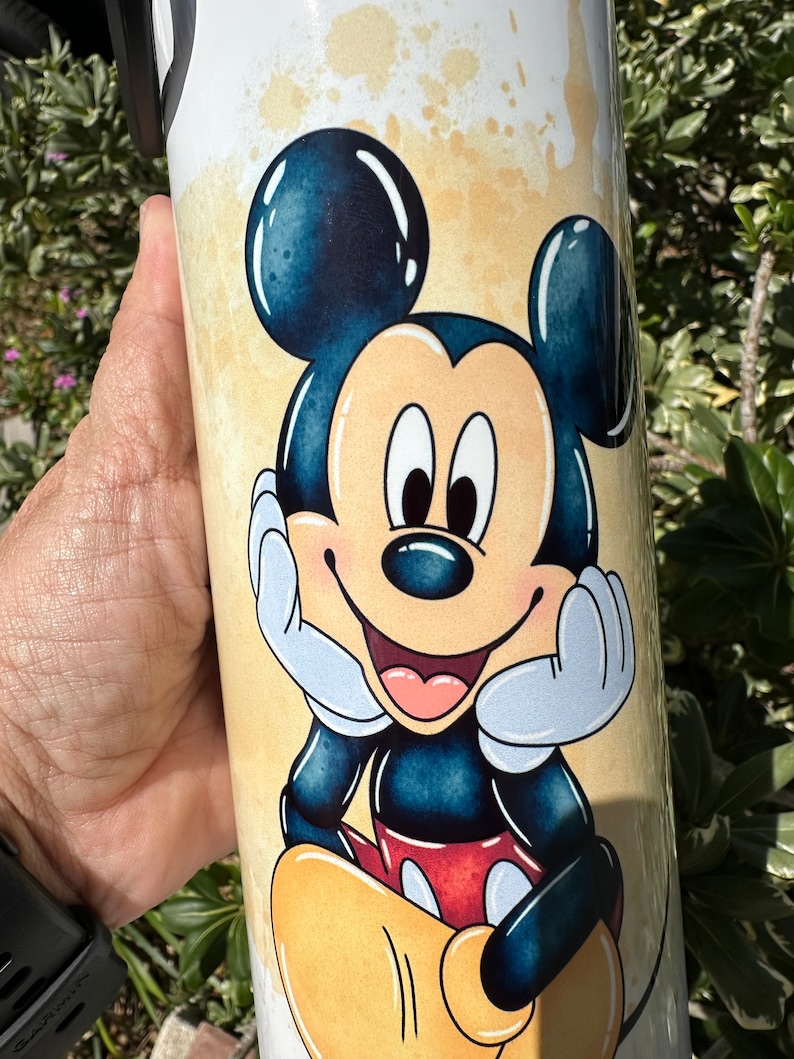 May include: A white tumbler with a cartoon image of Mickey Mouse. Mickey is smiling and has his hands on his chin. The background is a light yellow with brown splatters.