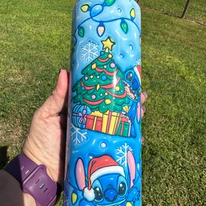 Może przedstawiać: Niebieski kubek termiczny ze stali nierdzewnej z świątecznym wzorem przedstawiającym Stitch'a z Lilo & Stitch. Wz&oacute;r zawiera choinkę, prezenty i płatki śniegu. Stitch ma na sobie czapkę Świętego Mikołaja.