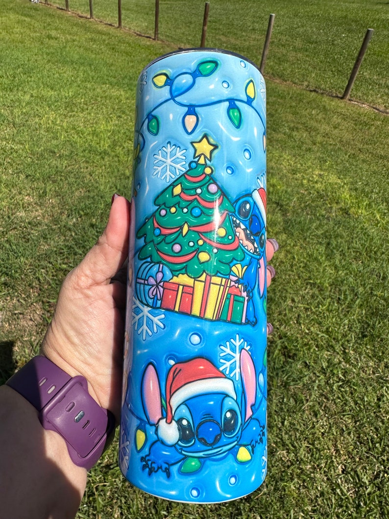 Może przedstawiać: Niebieski kubek termiczny ze stali nierdzewnej z świątecznym wzorem przedstawiającym Stitch'a z Lilo & Stitch. Wz&oacute;r zawiera choinkę, prezenty i płatki śniegu. Stitch ma na sobie czapkę Świętego Mikołaja.