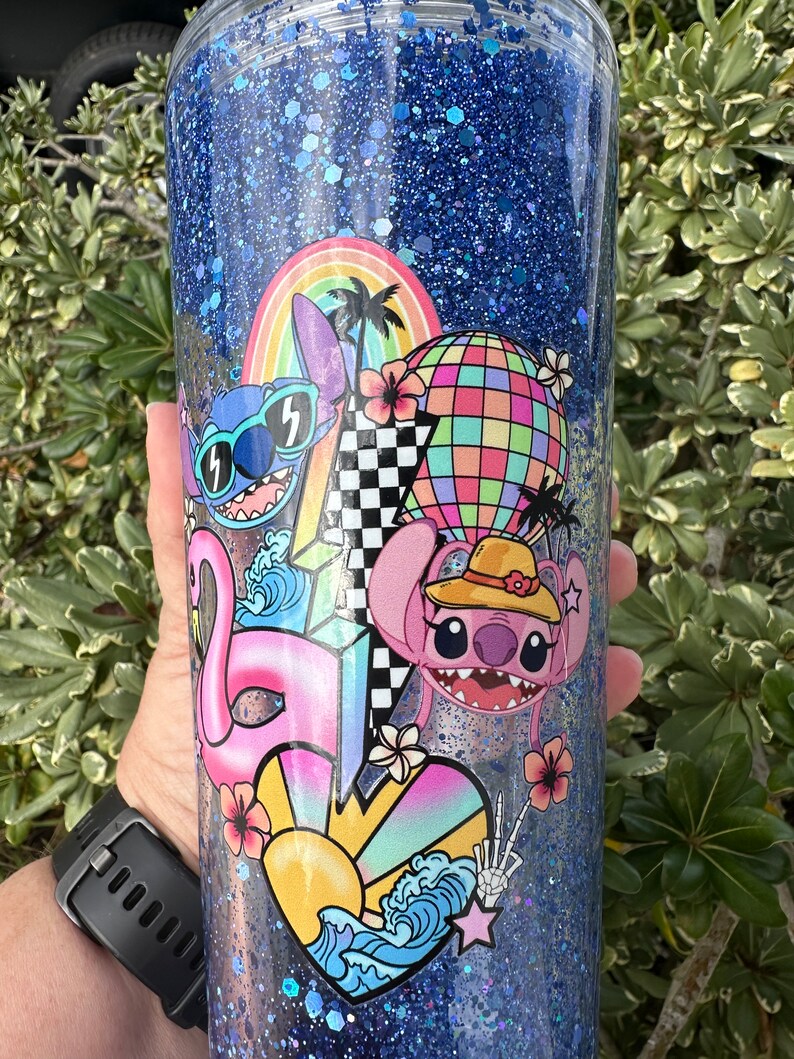 K&ouml;nnte beinhalten: Ein blauer Glitzerbecher mit einem farbenfrohen Design mit Stitch und Angel aus Lilo & Stitch. Das Design beinhaltet einen Regenbogen, Palmen, eine Discokugel, einen Flamingo, ein Herz, eine Sonne und Wellen.