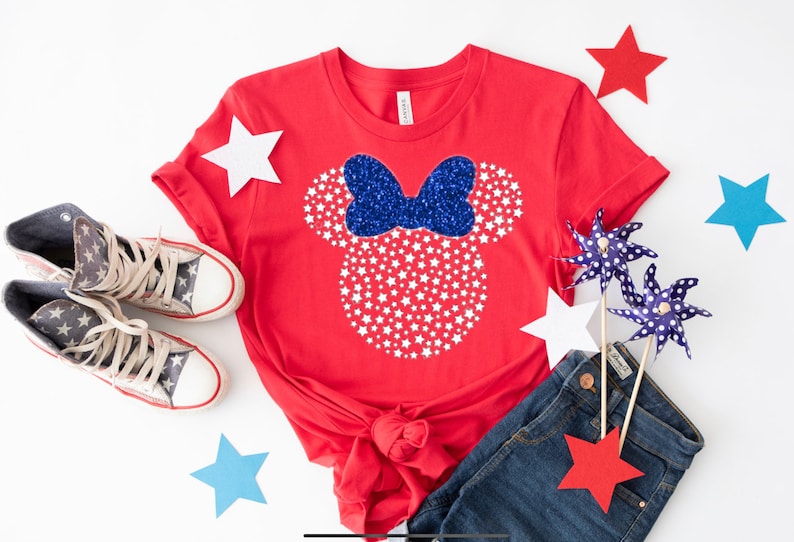 May include: Red t-shirt with a white and blue glitter Mickey Mouse head design. The design features a blue bow and white stars. The shirt is paired with blue jeans and red, white, and blue accessories.