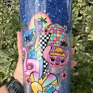 K&ouml;nnte beinhalten: Ein blauer Glitzerbecher mit einem farbenfrohen Design mit Stitch und Angel aus Lilo & Stitch. Das Design beinhaltet einen Regenbogen, Palmen, eine Discokugel, einen Flamingo und ein Herz mit einer Sonne und Wellen.