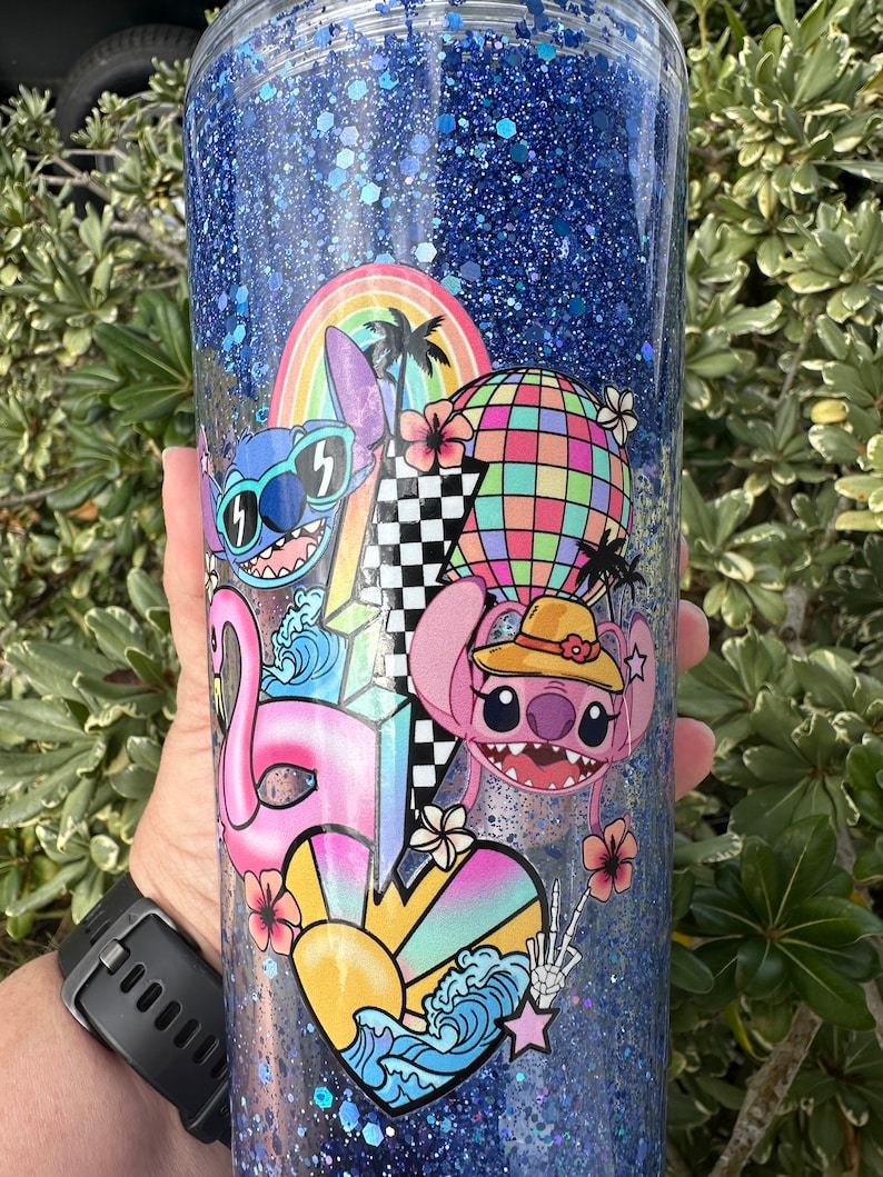 K&ouml;nnte beinhalten: Ein blauer Glitzerbecher mit einem farbenfrohen Design mit Stitch und Angel aus Lilo & Stitch. Das Design beinhaltet einen Regenbogen, Palmen, eine Discokugel, einen Flamingo und ein Herz mit einer Sonne und Wellen.