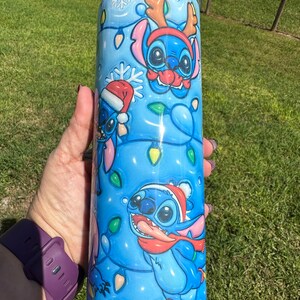 Może przedstawiać: Niebieski kubek ze stali nierdzewnej z świątecznym wzorem przedstawiającym Stitch'a z Lilo & Stitch. Wz&oacute;r przedstawia Stitch'a w czapce Świętego Mikołaja, czapce renifera i szaliku. Tło jest niebieskie z białymi płatkami śniegu i świątecznymi lampkami.