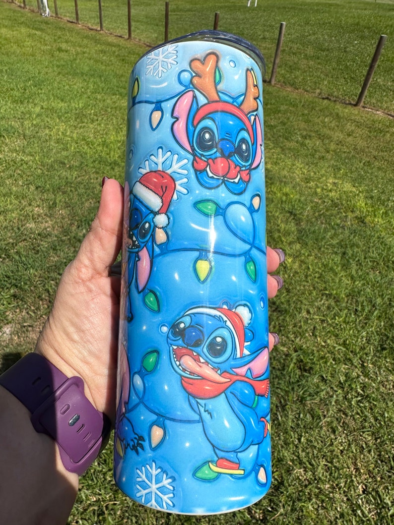 Może przedstawiać: Niebieski kubek ze stali nierdzewnej z świątecznym wzorem przedstawiającym Stitch'a z Lilo & Stitch. Wz&oacute;r przedstawia Stitch'a w czapce Świętego Mikołaja, czapce renifera i szaliku. Tło jest niebieskie z białymi płatkami śniegu i świątecznymi lampkami.
