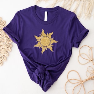 May include: A purple t-shirt featuring a gold glitter sun design. The short-sleeved shirt is knotted at the waist. The sun design is centered on the front of the shirt.
