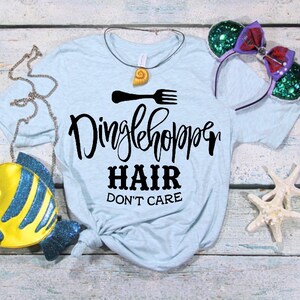 Może przedstawiać: Jasnoniebieska koszulka z napisem "Dinglehopper Hair Don't Care" w kolorze czarnym. Koszulka jest zawiązana na dole. Na zdjęciu widać r&oacute;wnież ż&oacute;łtą torebkę w kształcie ryby, naszyjnik z muszli i zielono-fioletową opaskę na głowę.