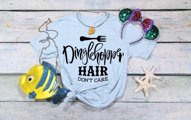 Może przedstawiać: Jasnoniebieska koszulka z napisem "Dinglehopper Hair Don't Care" w kolorze czarnym. Koszulka jest zawiązana na dole. Na zdjęciu widać r&oacute;wnież ż&oacute;łtą torebkę w kształcie ryby, naszyjnik z muszli i zielono-fioletową opaskę na głowę.
