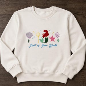 May include: A cream-colored crewneck sweatshirt featuring an embroidered design of a mermaid with red hair and a green tail, a yellow trident, a purple seashell, a pink starfish, and coral. The text "Part of Your World" is embroidered in blue.