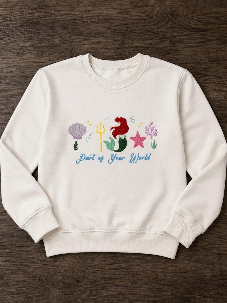 May include: A cream-colored crewneck sweatshirt featuring an embroidered design of a mermaid with red hair and a green tail, a yellow trident, a purple seashell, a pink starfish, and coral. The text "Part of Your World" is embroidered in blue.
