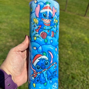 Może przedstawiać: Niebieski i biały kubek ze stali nierdzewnej z świątecznym wzorem przedstawiającym Stitch'a z Lilo & Stitch'a w czapce Świętego Mikołaja trzymającego świąteczne lampki.