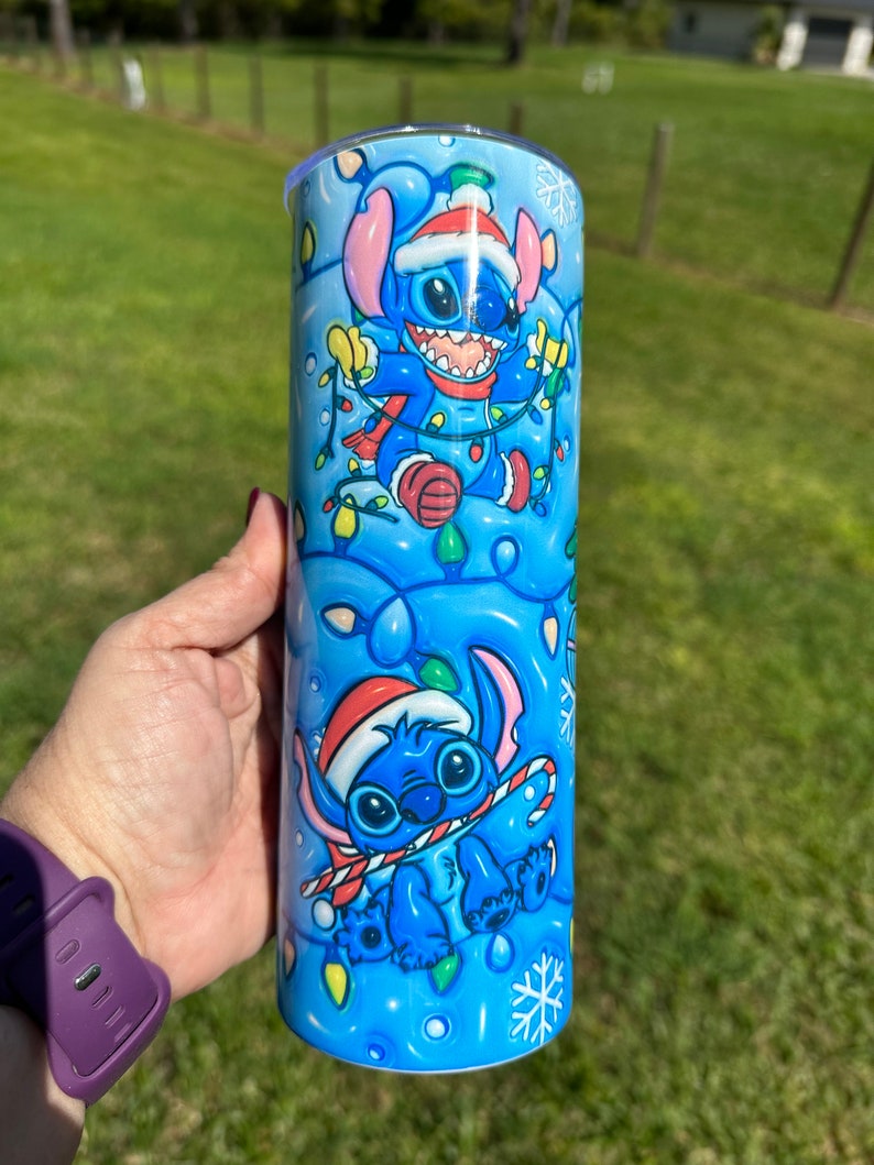 Może przedstawiać: Niebieski i biały kubek ze stali nierdzewnej z świątecznym wzorem przedstawiającym Stitch'a z Lilo & Stitch'a w czapce Świętego Mikołaja trzymającego świąteczne lampki.
