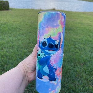 Puede incluir: Un vaso de acero inoxidable azul y blanco con un dise&ntilde;o tie-dye y una imagen de dibujos animados de Stitch de Lilo & Stitch sosteniendo un juguete de peluche.