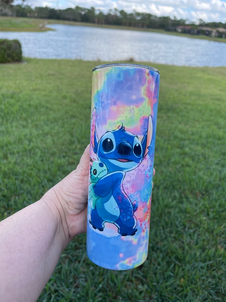 Puede incluir: Un vaso de acero inoxidable azul y blanco con un dise&ntilde;o tie-dye y una imagen de dibujos animados de Stitch de Lilo & Stitch sosteniendo un juguete de peluche.