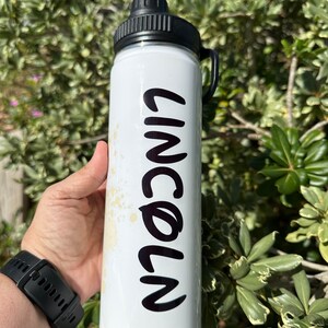 May include: White water bottle with a black lid and handle. The bottle has the name "LINCOLN" written in black on the side.