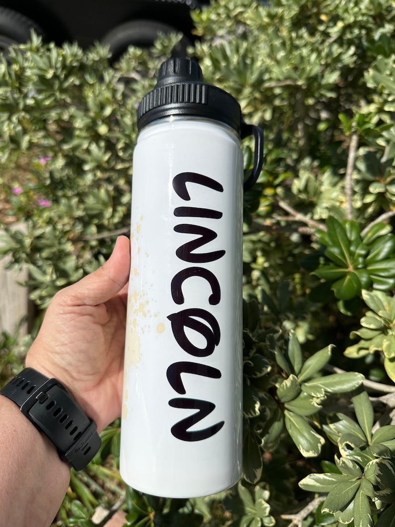 May include: White water bottle with a black lid and handle. The bottle has the name "LINCOLN" written in black on the side.