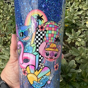 K&ouml;nnte beinhalten: Ein blauer Glitzerbecher mit einem farbenfrohen Design mit Stitch aus Lilo & Stitch. Das Design beinhaltet einen Regenbogen, Palmen, einen Hei&szlig;luftballon, einen Flamingo und ein Herz mit Wellen.