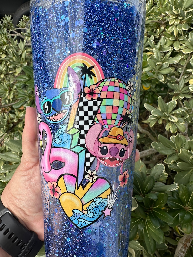 K&ouml;nnte beinhalten: Ein blauer Glitzerbecher mit einem farbenfrohen Design mit Stitch aus Lilo & Stitch. Das Design beinhaltet einen Regenbogen, Palmen, einen Hei&szlig;luftballon, einen Flamingo und ein Herz mit Wellen.