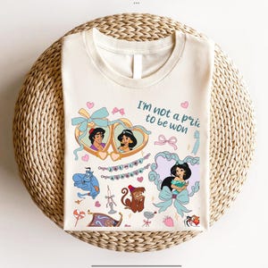 May include: A white t-shirt with a graphic print of Jasmine and Aladdin from Disney's Aladdin. The graphic includes a blue genie, a monkey, and the text "I'm not a prize to be won".