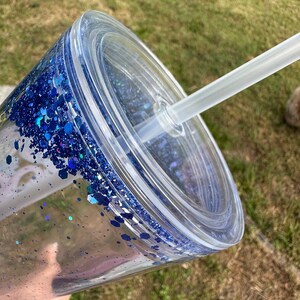 K&ouml;nnte beinhalten: Ein durchsichtiger Plastikbecher mit einem blauen Glitzerdesign und einem durchsichtigen Deckel. Ein durchsichtiger Plastikstrohhalm ist durch den Deckel gesteckt.