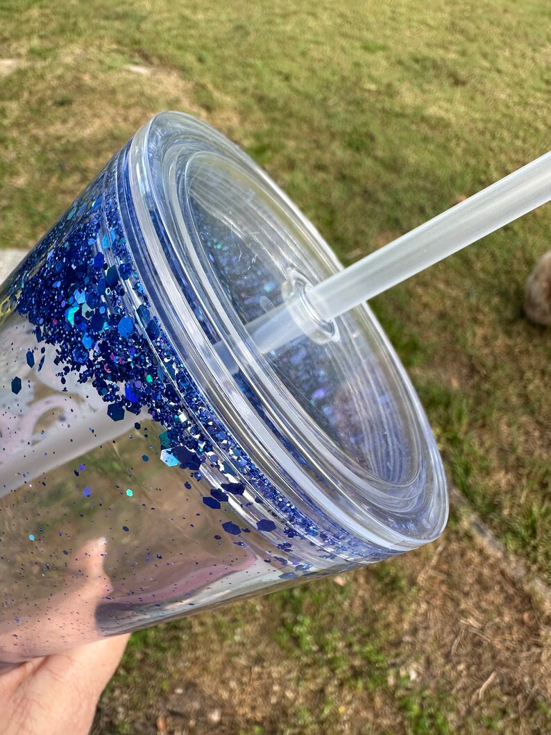 K&ouml;nnte beinhalten: Ein durchsichtiger Plastikbecher mit einem blauen Glitzerdesign und einem durchsichtigen Deckel. Ein durchsichtiger Plastikstrohhalm ist durch den Deckel gesteckt.