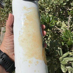 May include: White stainless steel water bottle with a black lid and handle. The bottle has a yellow stain on the side.