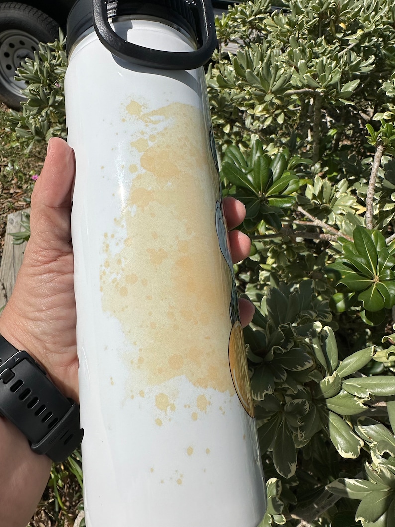 May include: White stainless steel water bottle with a black lid and handle. The bottle has a yellow stain on the side.
